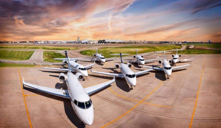 Seven private jets parked on an airport tarmac under a vibrant, cloudy sunset sky.