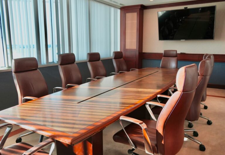 A sunlit conference room with a long wooden table, surrounded by brown leather chairs, and a wall-mounted TV.