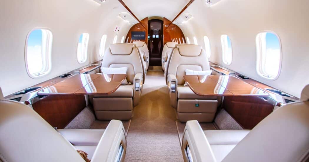 Interior of a luxurious private jet with cream leather seats, polished wooden tables, and large windows on both sides.