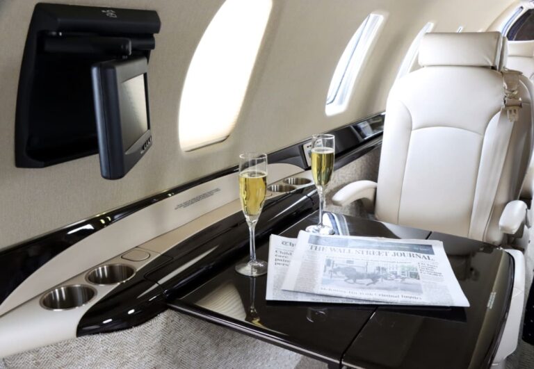 Luxurious private jet interior with champagne glasses and a newspaper on a glossy table beside leather seating.