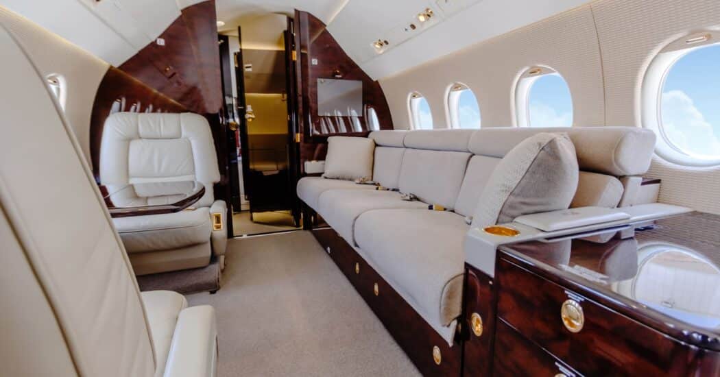 Interior of a private jet with plush leather seats, a beige sofa, wooden panels, and oval windows showing a view of clouds outside.