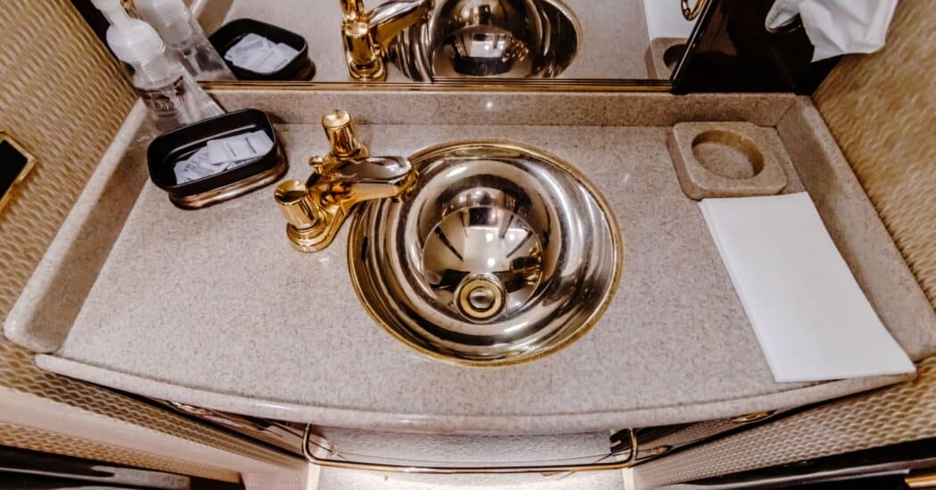 Lavish airplane bathroom sink with a gold faucet and basin, a water bottle, tissue box, and a folded towel on the marble countertop.