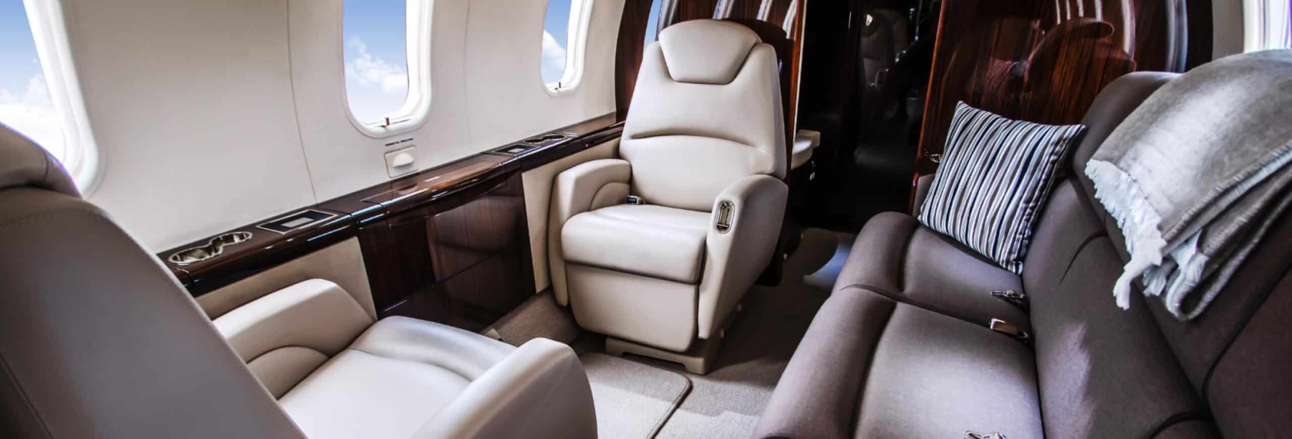 Luxurious private jet interior with plush beige leather seats and a gray cushioned sofa, featuring wood paneling and large windows.