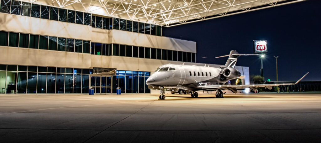 Private jet parked outside a modern airport terminal at night, under a brightly lit canopy with a Phillips 66 sign in the background.