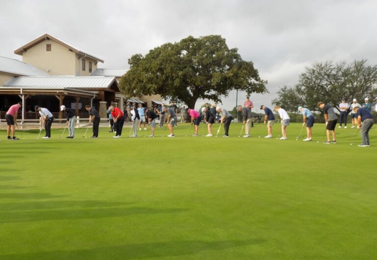 A group of people lined up on a golf green, practicing putting near a clubhouse under a cloudy sky.