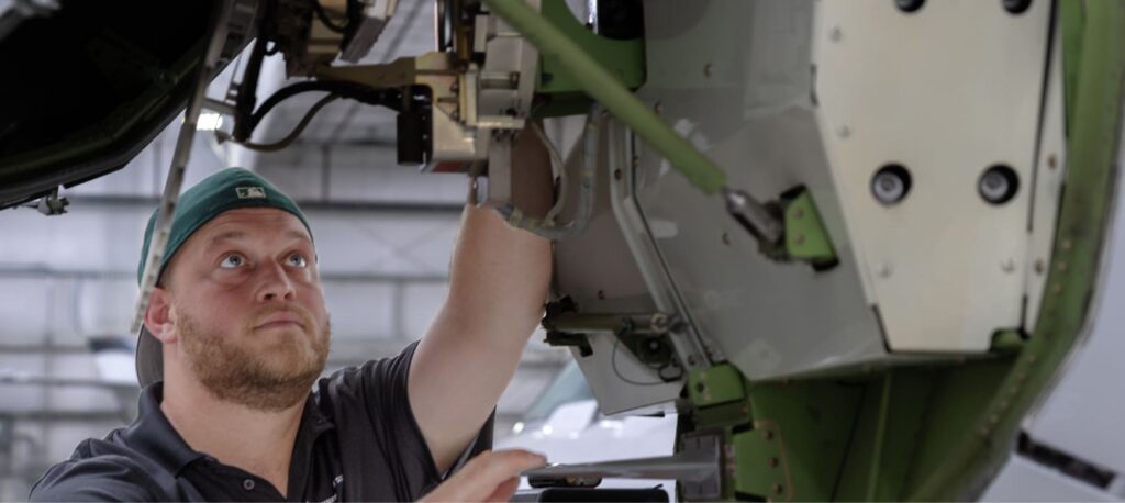 A person in a cap inspects machinery inside an aircraft hangar, surrounded by metal components.