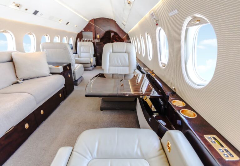 Interior of a luxurious private jet cabin with plush beige leather seating, wood accents, and multiple windows with views of the sky.