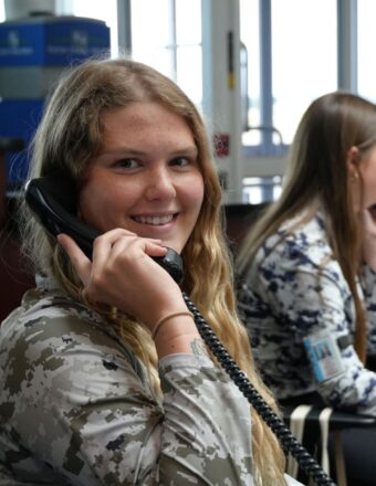 Two women are sitting and talking on phones. The woman in the foreground is smiling and has long, wavy hair.